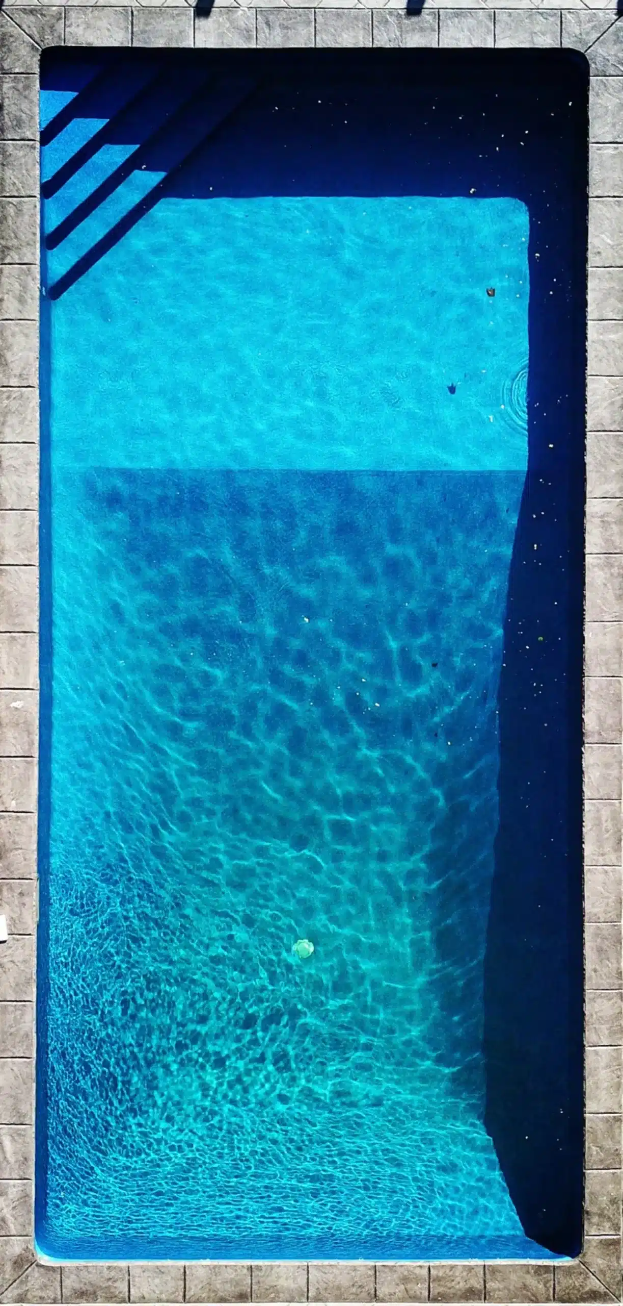 Rectangular pool with clear blue water, ripples from sunlight, partial shadows, stairs at top left, and a few floating objects.