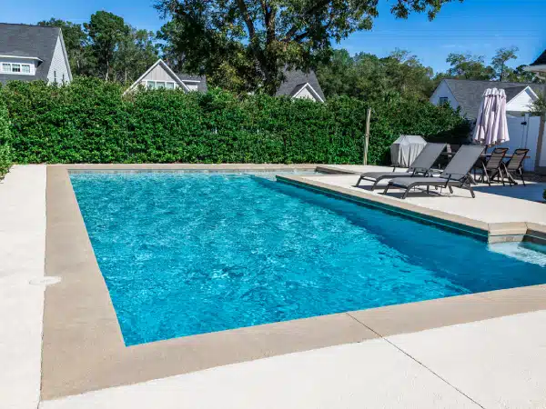 Backyard pool with clear blue water, concrete deck, lounge chairs with umbrellas, green shrubs, and houses in the background.