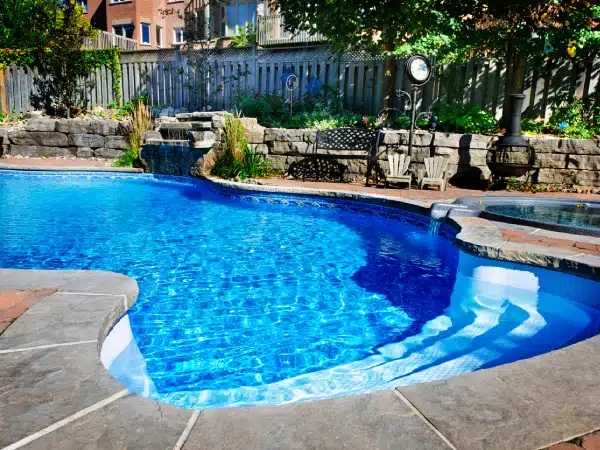 Backyard pool with clear blue water, stone patio, steps, landscaped plants, and a bench by a sunlit fence.