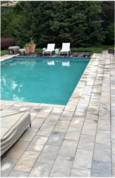 Rectangular backyard pool with stone tile, two white lounge chairs by greenery, and a cushioned lounger in front.