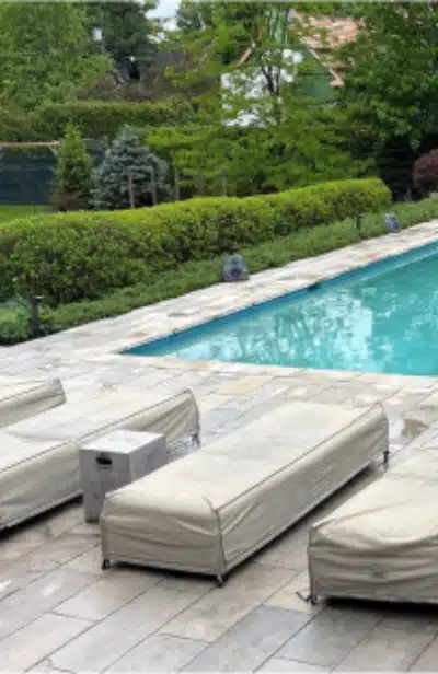 Four lounge chairs with covers beside a rectangular pool, stone tile deck, and lush green bushes and trees in the background.