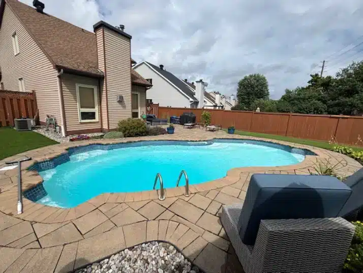 Irregular in-ground pool in a backyard with stone patio, lounge chairs, wooden fence, greenery, and cloudy sky.