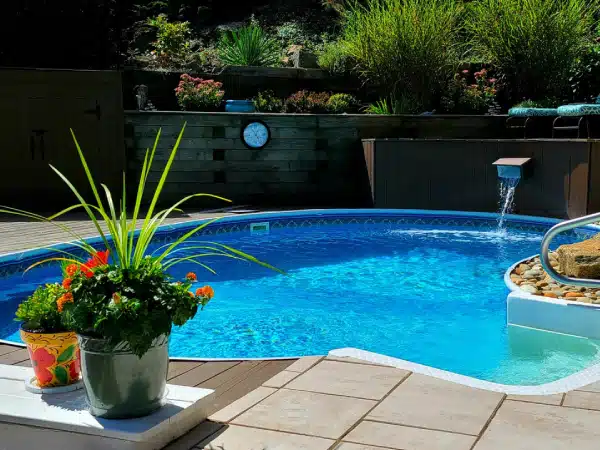 Backyard pool with clear blue water, a waterfall feature, curved handrail, potted plants, greenery, and a fence clock.