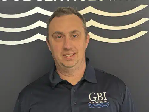 Certified GBI Team member in navy shirt standing against dark wall with white wave patterns.