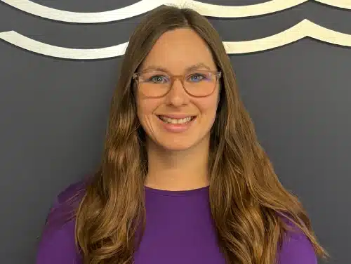 Smiling woman with long brown hair, glasses, and purple shirt against a dark gray wall.